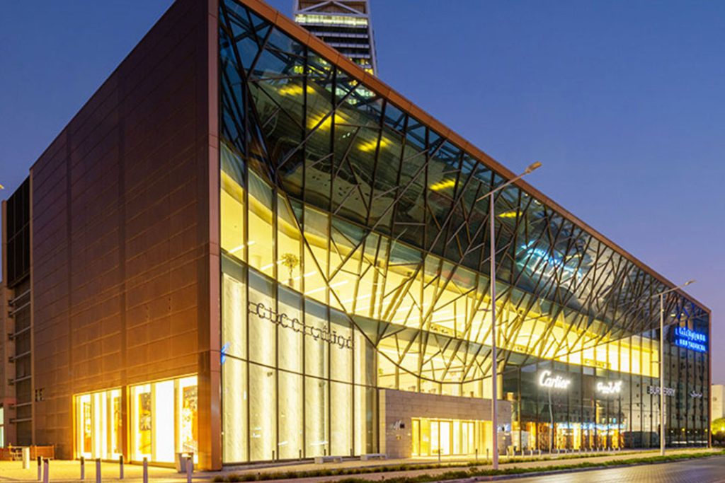 Al Faisaliyah Shopping Centre at dusk: lit faceted glass facade with Cartier and Gucci shopfronts