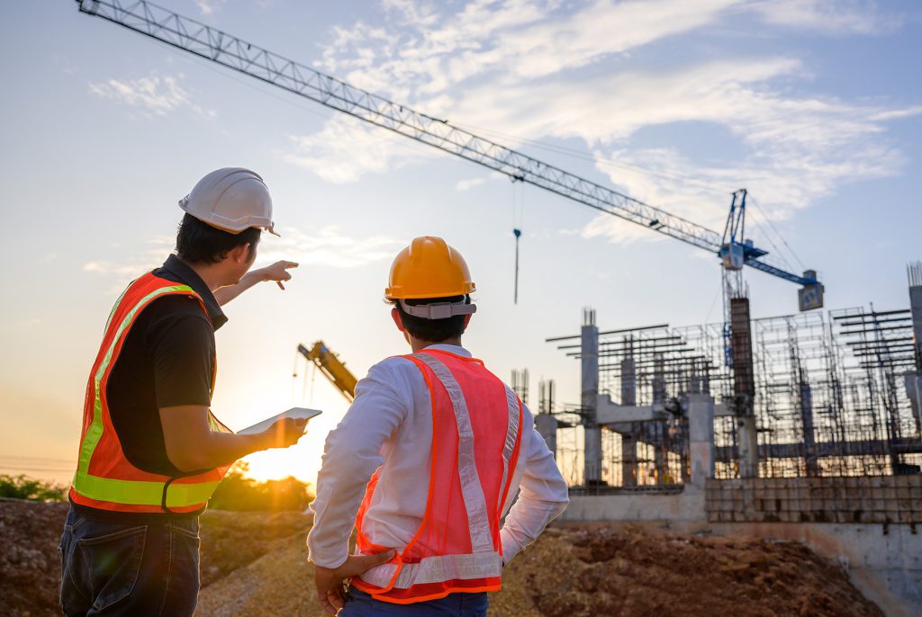 Two engineers in hard hats and hi-vis vests looking towards a crane and concrete frame at sunset