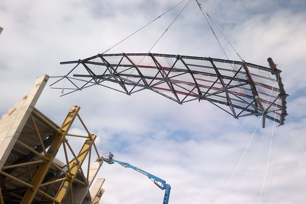 Steel roof truss suspended from crane cables above the OVO Hydro, with a worker on a boom lift