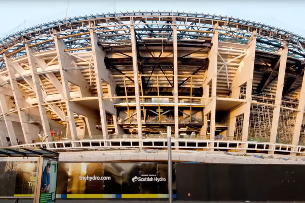 OVO Hydro under construction: concrete frame and roof steelwork behind hoarding reading 'thehydro.com' and 'Scottish Hydro'